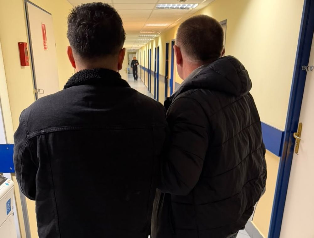 Dwóch mężczyzn w ciemnych kurtkach stoi na korytarzu i patrzy w stronę osoby oddalonej w głębi.