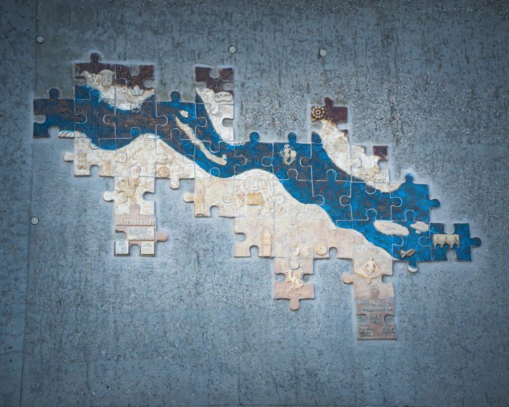 Ceramiczna mozaika złożona z puzzli, przedstawiająca niebieską rzekę na jasnym tle, zamontowana na szarej ścianie.