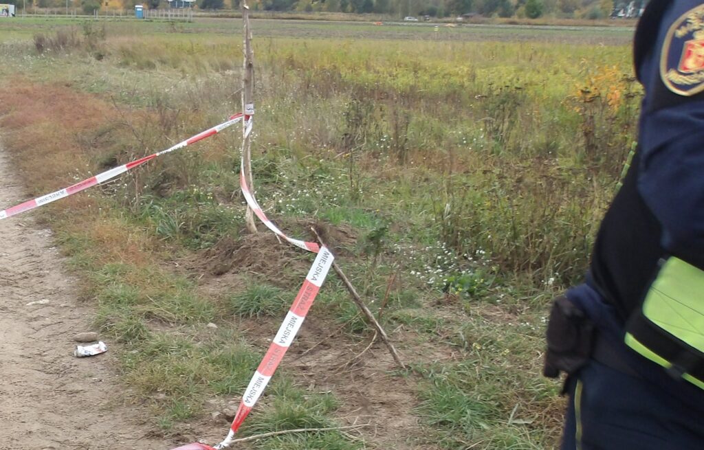 Teren przy polu odgrodzony taśmą ostrzegawczą, obok stoi funkcjonariusz w mundurze; na ziemi leży niewielki przedmiot.