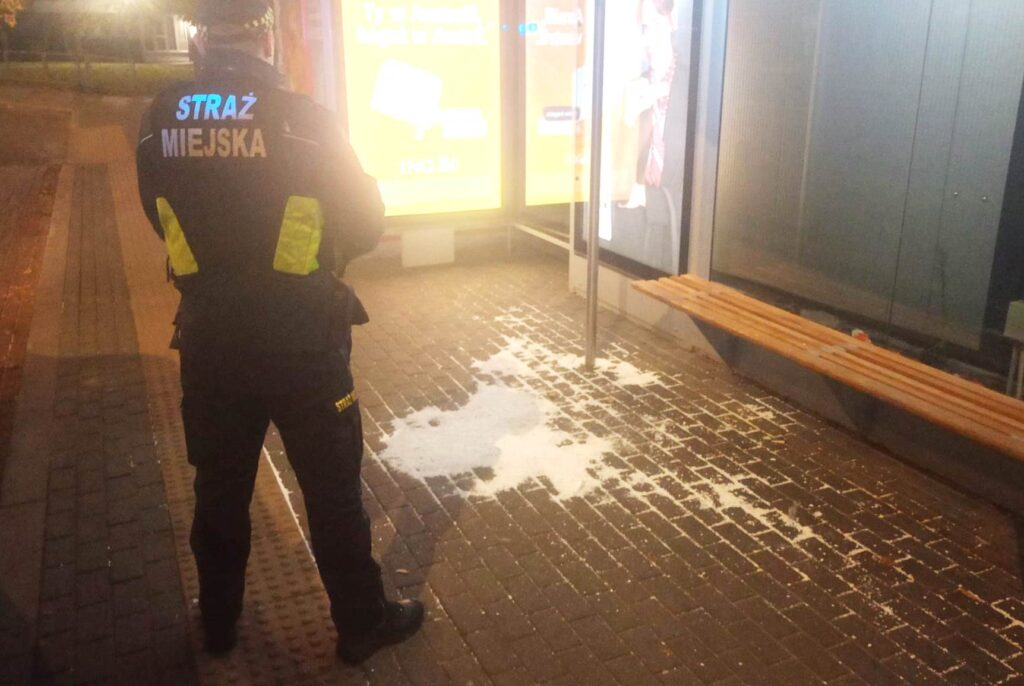Strażnik miejski stoi na chodniku obok ławki i rozrzuconych śmieci nocą.