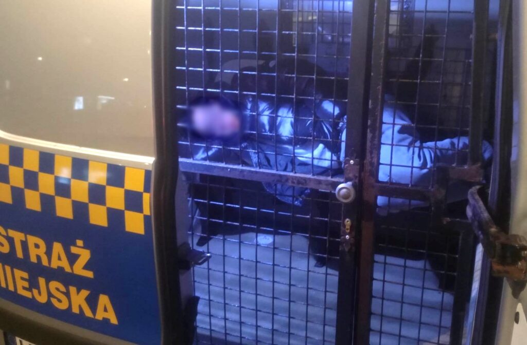 Osoba siedzi za kratami w przedziale pojazdu straży miejskiej, z rozmytą twarzą.