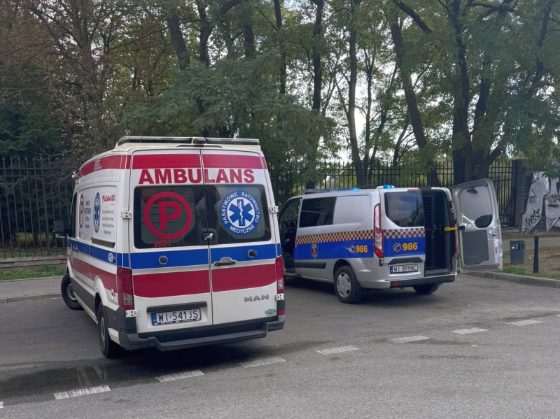 Dwie karetki zaparkowane przy ogrodzeniu pod drzewami; tylne drzwi jednego ambulansu są otwarte.