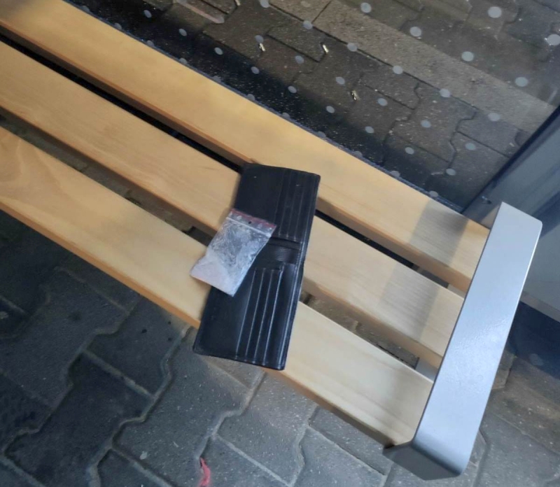 Czarny portfel z małą przezroczystą torebką leży na drewnianej ławce przy przeszklonej ścianie.