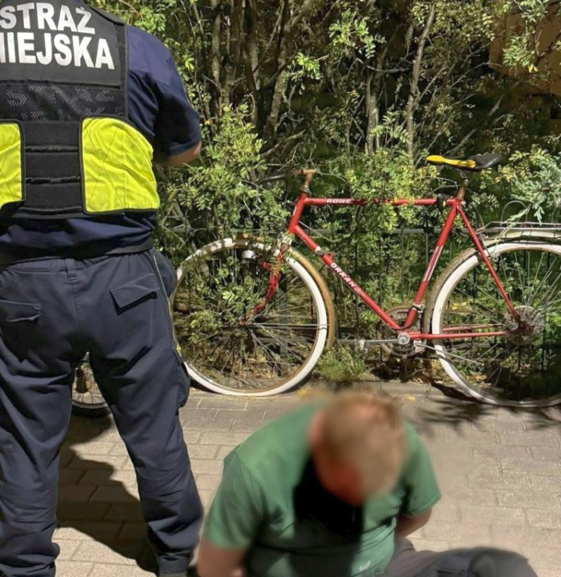 Funkcjonariusz straży miejskiej stoi obok siedzącego na chodniku mężczyzny; za nimi stoi czerwony rower przy krzewach.