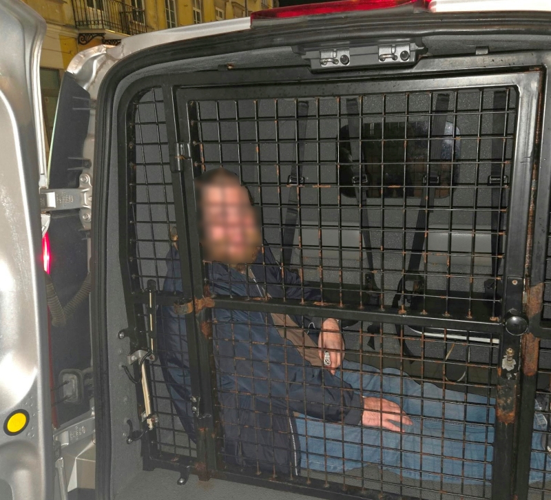 Mężczyzna siedzi za kratą w przedziale transportowym pojazdu, z twarzą zasłoniętą pikselami.