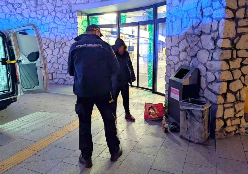 Strażnik miejski rozmawia z osobą w kapturze przy wejściu do budynku, obok zaparkowanego radiowozu.