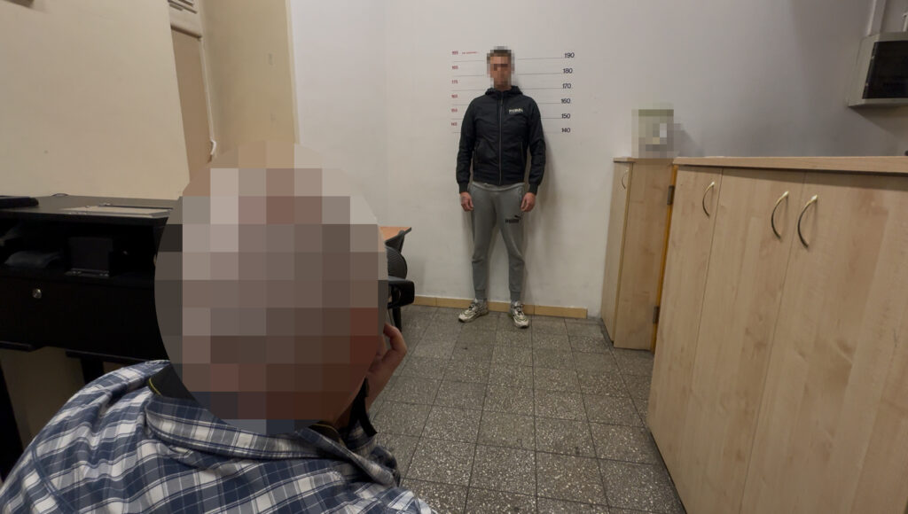Mężczyzna stoi przy ścianie ze skalą wzrostu, a drugi siedzi na pierwszym planie w pomieszczeniu.