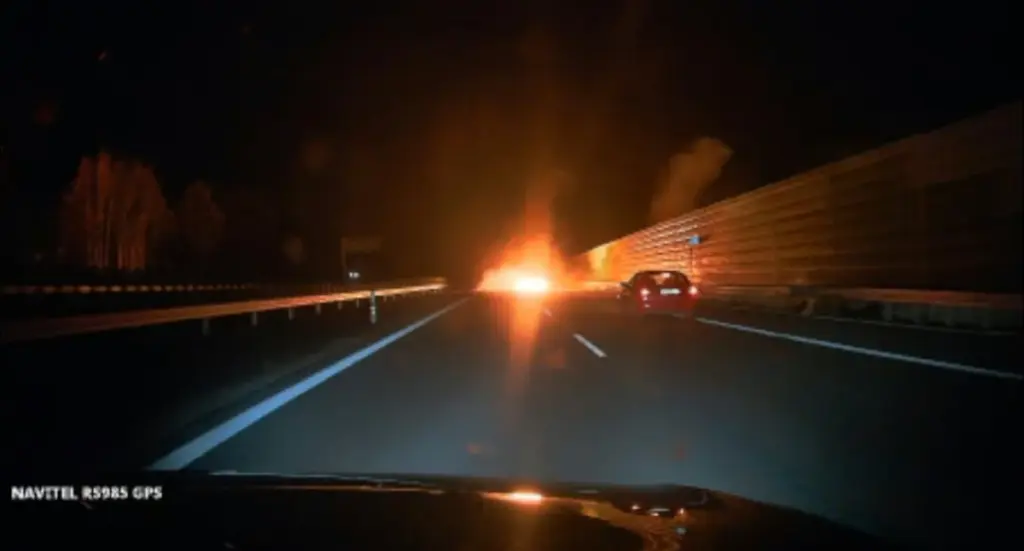 Płonący pojazd na nocnej autostradzie, obok jedzie samochód osobowy; ujęcie z kamery samochodowej.