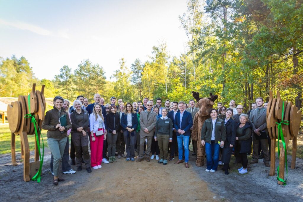 Grupa osób pozuje przy drewnianej bramie z figurą łosia, na leśnej polanie otoczonej jesiennymi drzewami.