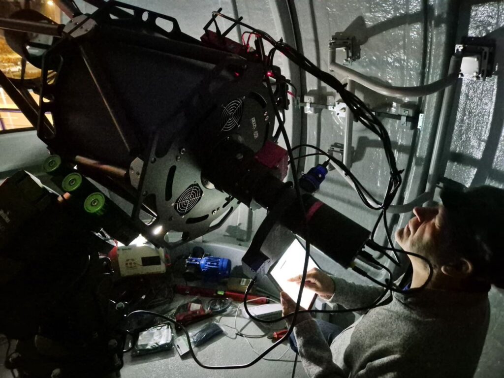 Mężczyzna obsługuje sprzęt filmowy z kamerą i kablami w ciemnym pomieszczeniu technicznym.