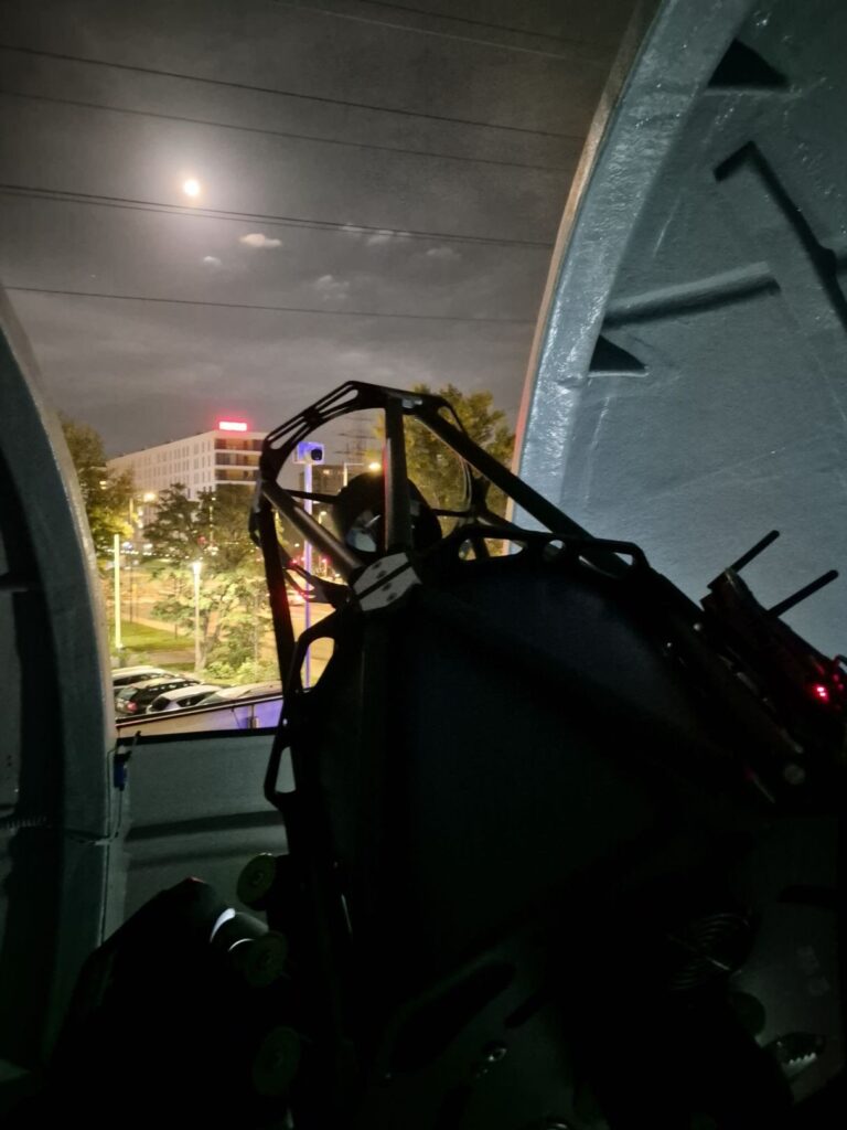 Duży teleskop astronomiczny ustawiony wewnątrz kopuły obserwatorium nocą, z widokiem na oświetlone budynki i ulicę.