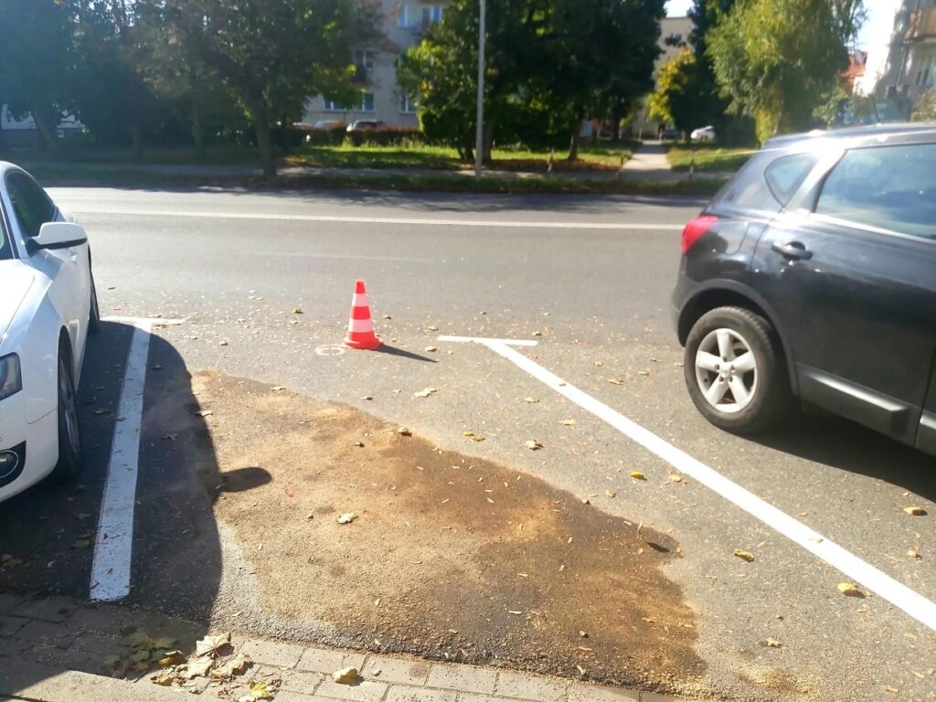 Pomarańczowy pachołek stoi na parkingu między białym i ciemnym samochodem, obok zabrudzonej nawierzchni.