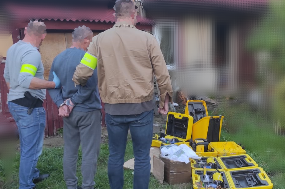 Policjanci prowadzą zatrzymanego mężczyznę obok rozłożonych żółtych urządzeń i walizek ze sprzętem na trawniku