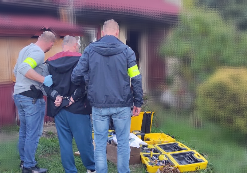 Funkcjonariusze prowadzą zatrzymanego mężczyznę obok żółtych walizek ze sprzętem przy budynku.