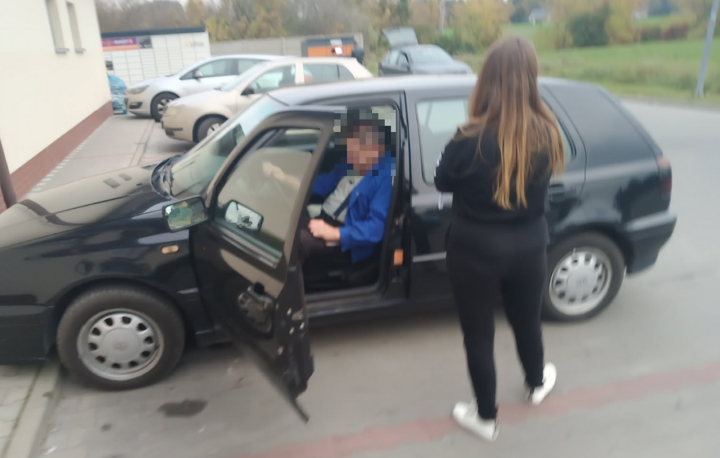Mężczyzna siedzi w otwartym samochodzie, a kobieta stoi obok na parkingu przy budynku.