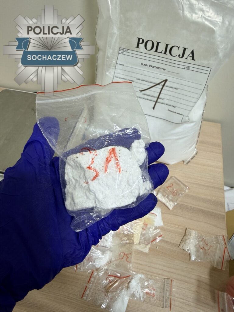 Ręka w niebieskiej rękawiczce trzyma woreczek z białą substancją, obok leżą zabezpieczone próbki i dokumenty policji.
