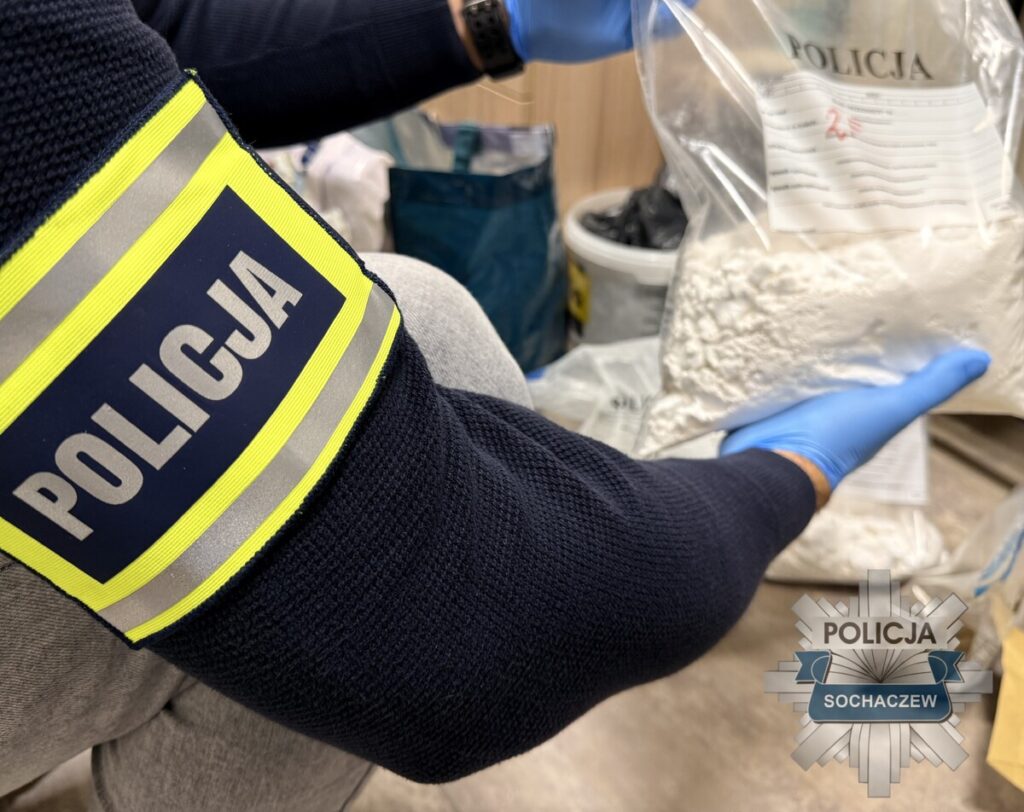 Policjant w niebieskiej rękawiczce trzyma przezroczysty worek z białym proszkiem, zabezpieczony przez policję.