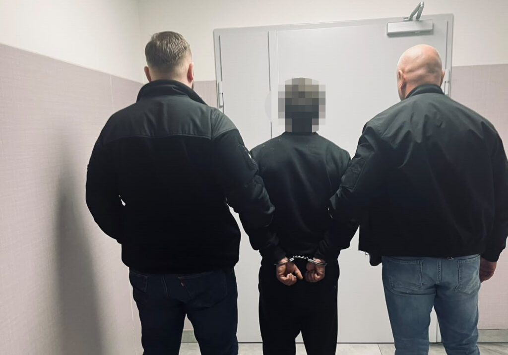 Dwóch funkcjonariuszy prowadzi zakutego w kajdanki mężczyznę korytarzem w stronę drzwi.