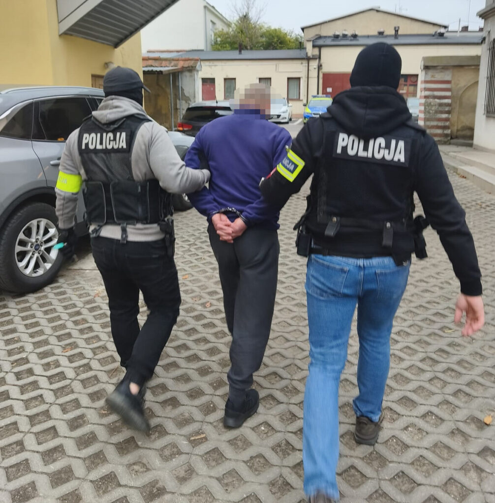 Dwóch policjantów prowadzi skutego kajdankami mężczyznę po podwórzu między budynkami i zaparkowanym samochodem.