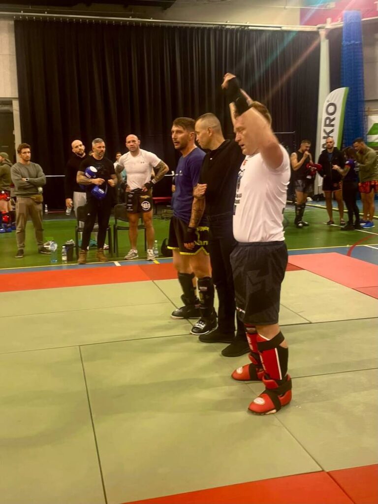 Mężczyzna w białej koszulce unosi rękę na macie do kickboxingu, obok stoją zawodnicy i sędzia.