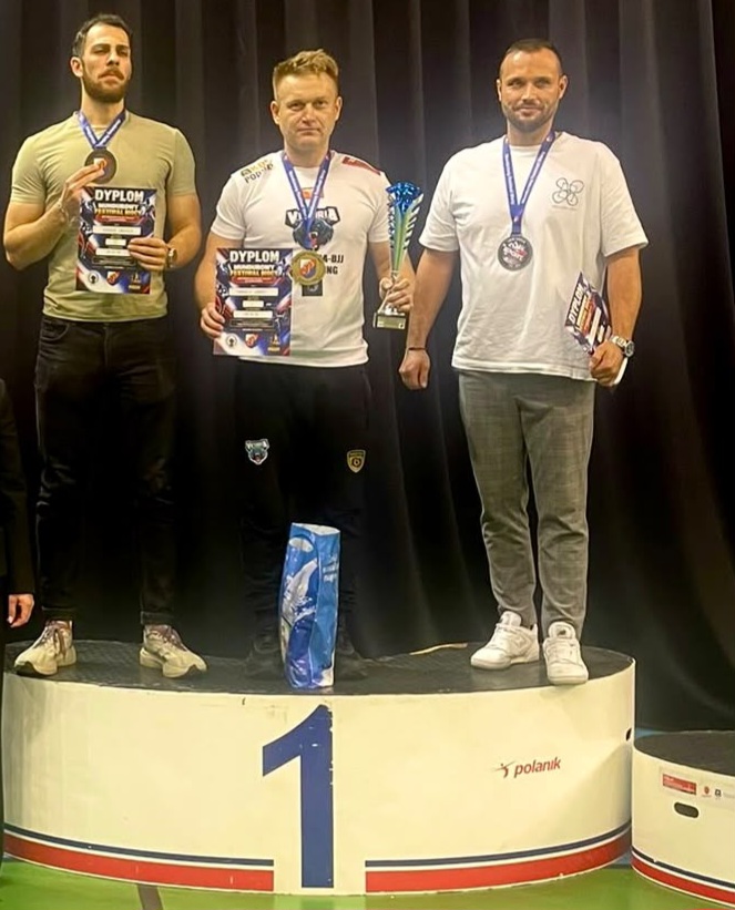 Trzej mężczyźni z medalami i dyplomami stoją na podium, a środkowy trzyma puchar.