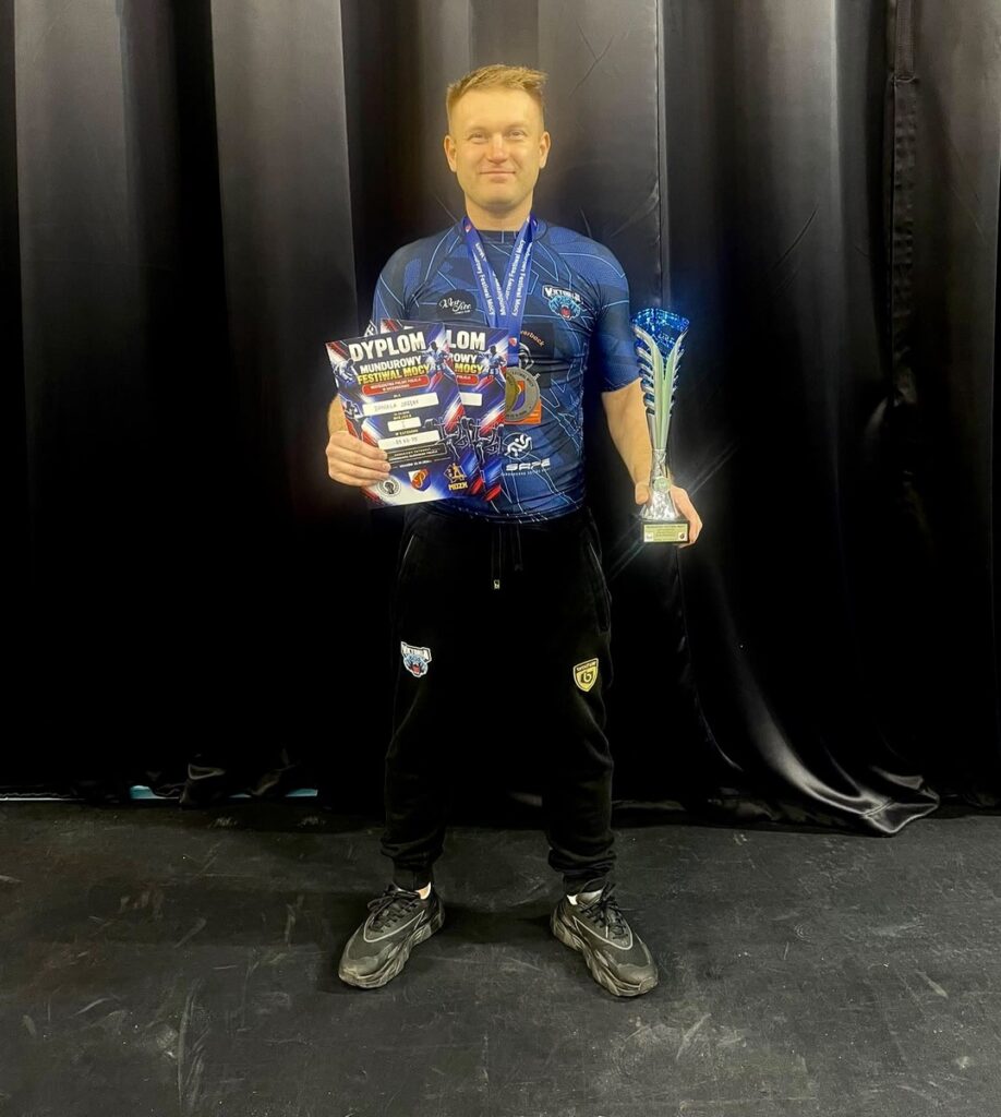 Mężczyzna w niebieskiej koszulce sportowej prezentuje dyplom i puchar, stojąc na tle czarnej kurtyny.