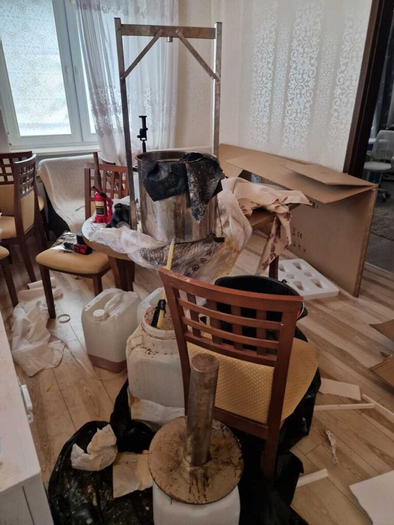 Metalowe urządzenia, kanistry i pojemniki stoją w zagraconym pokoju z drewnianymi krzesłami.