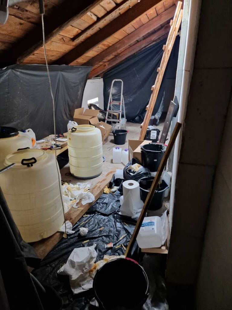 Zagracony strych z dużymi plastikowymi zbiornikami, kanistrami, wiadrami i drabiną.