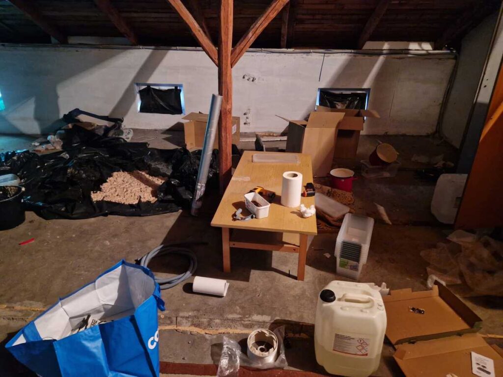 Zagracony strych z workami, kartonami, pojemnikami i stołem z drobnymi przedmiotami.