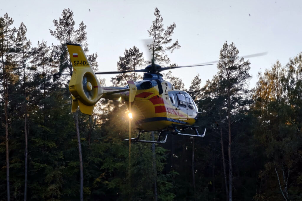 Żółto-czerwony helikopter leci nisko nad lasem, na tle drzew i zachodzącego słońca.