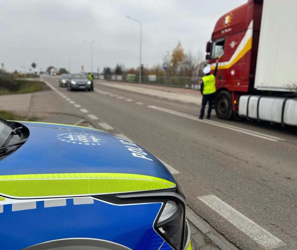 Policjant w kamizelce odblaskowej stoi przy czerwonej ciężarówce na drodze, obok widoczny radiowóz.