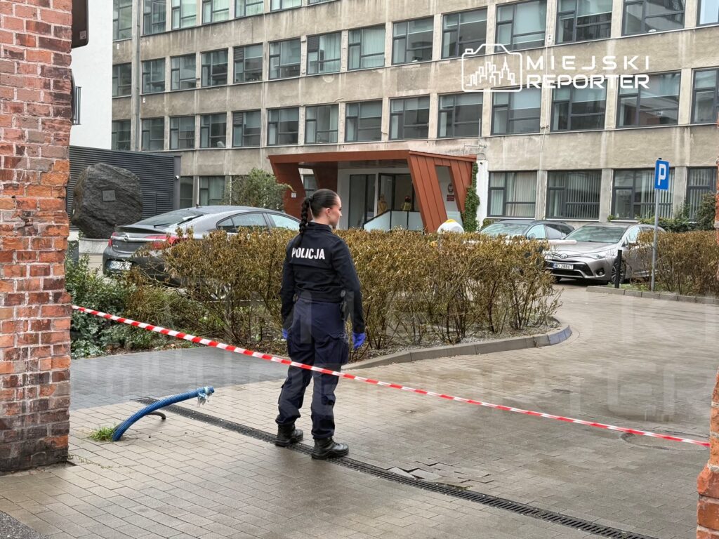 Policjant stoi za taśmą zabezpieczającą teren przed wejściem do biurowca.