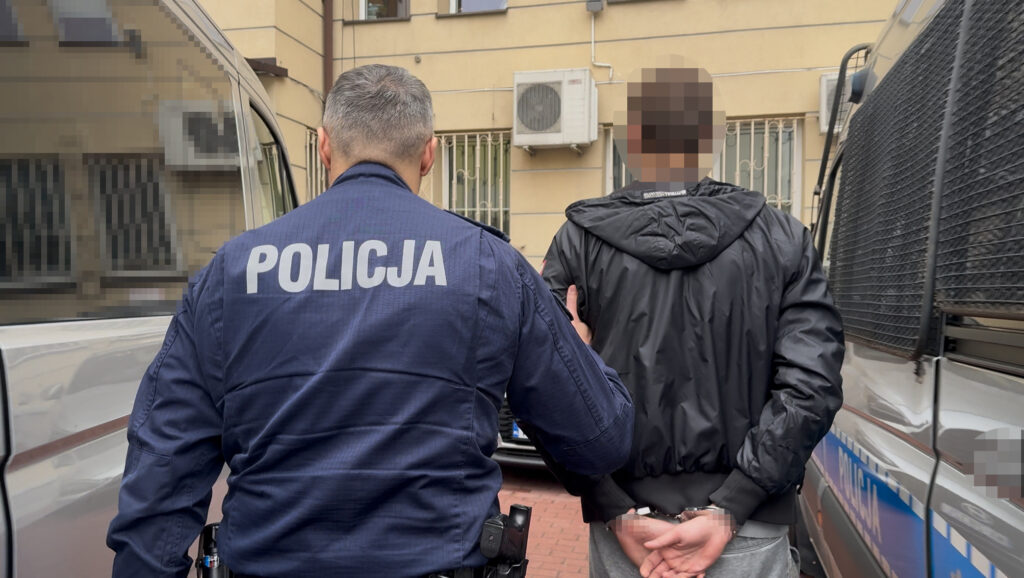 Policjant prowadzi skutego kajdankami mężczyznę między zaparkowanymi radiowozami na dziedzińcu budynku.