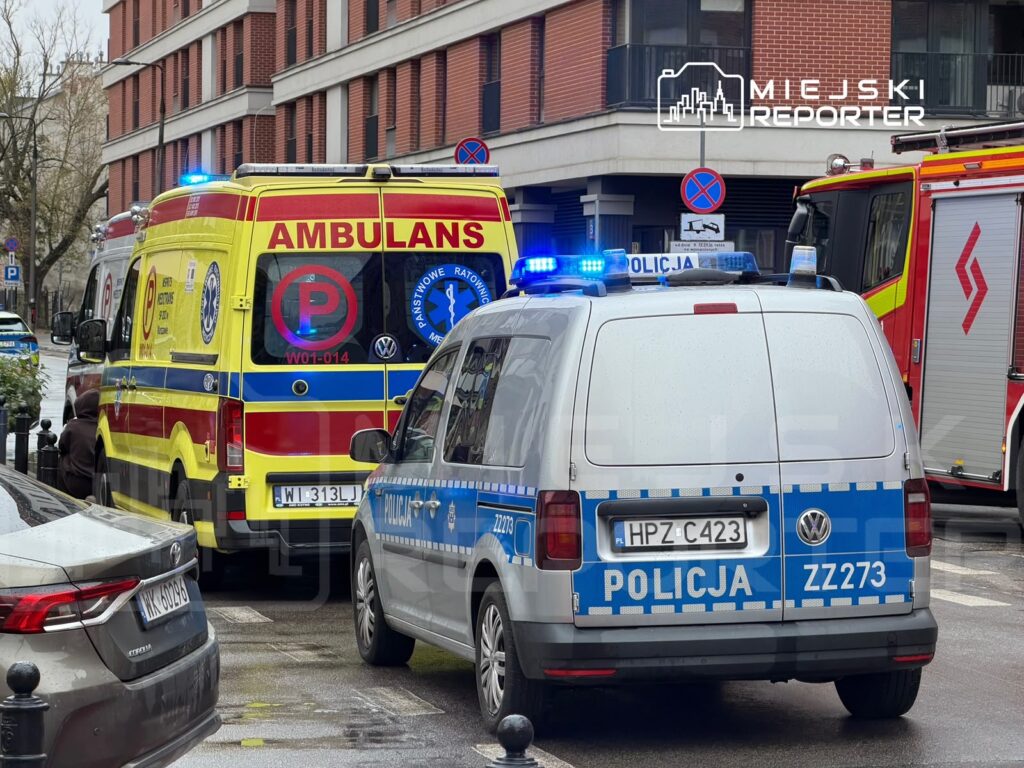 Karetka pogotowia, policyjny van i wóz strażacki stoją na ulicy przed budynkiem z czerwonej cegły.