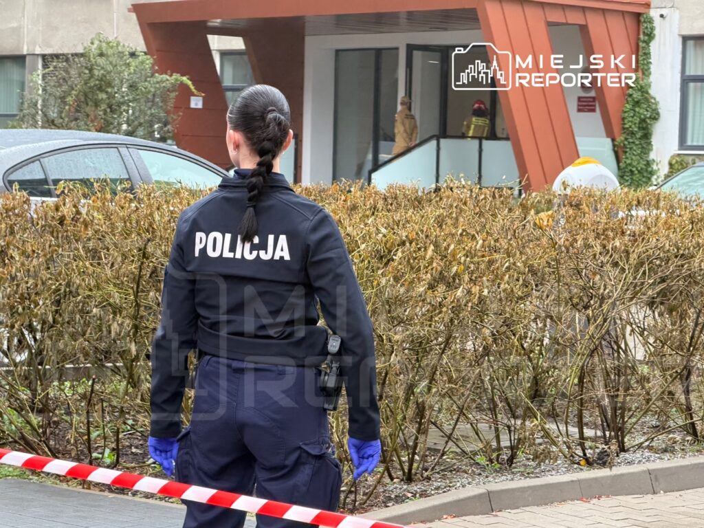 Funkcjonariuszka policji stoi za taśmą zabezpieczającą przed wejściem do budynku.