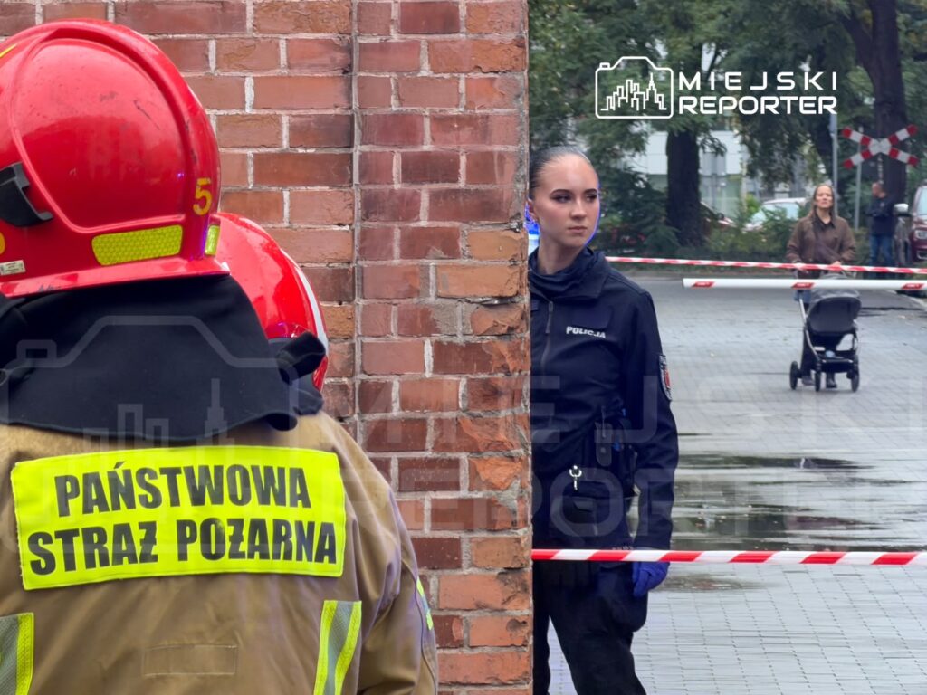Strażak w czerwonym hełmie stoi przy ceglanym murze, a policjantka zabezpiecza taśmą przejście obok wózka dziecięcego.