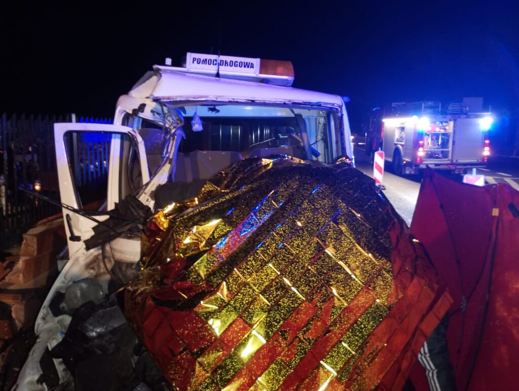 Uszkodzona kabina samochodu ciężarowego przykryta złotym kocem ratunkowym, obok wóz strażacki podczas nocnej akcji