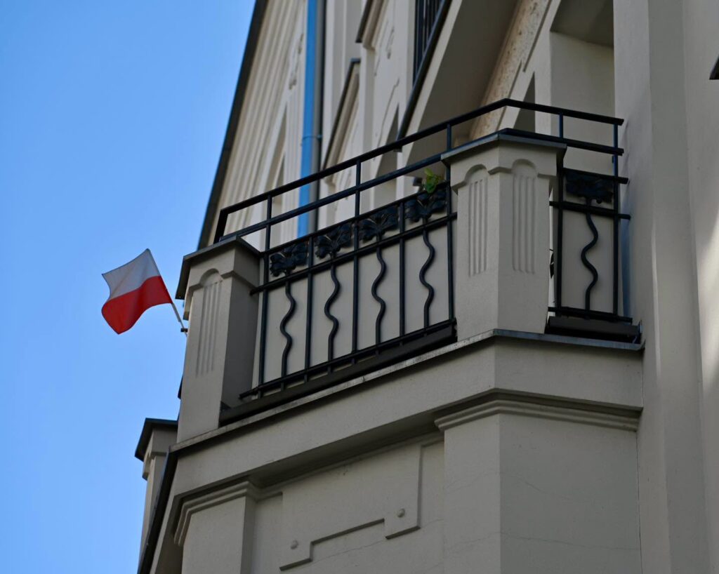 Polska flaga powiewa przy balkonie z czarną balustradą na elewacji jasnego budynku, na tle błękitnego nieba.