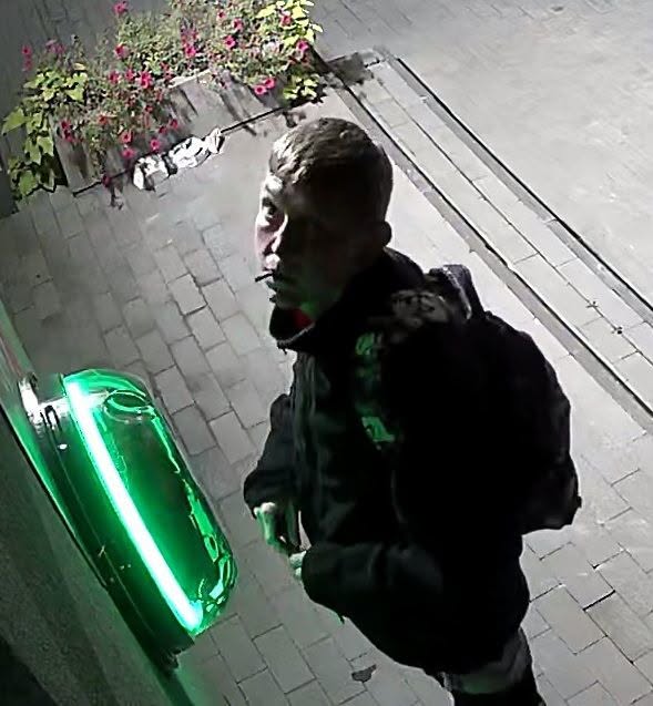 Mężczyzna w ciemnej kurtce i z plecakiem stoi obok podświetlonej zielonej skrzynki na chodniku.