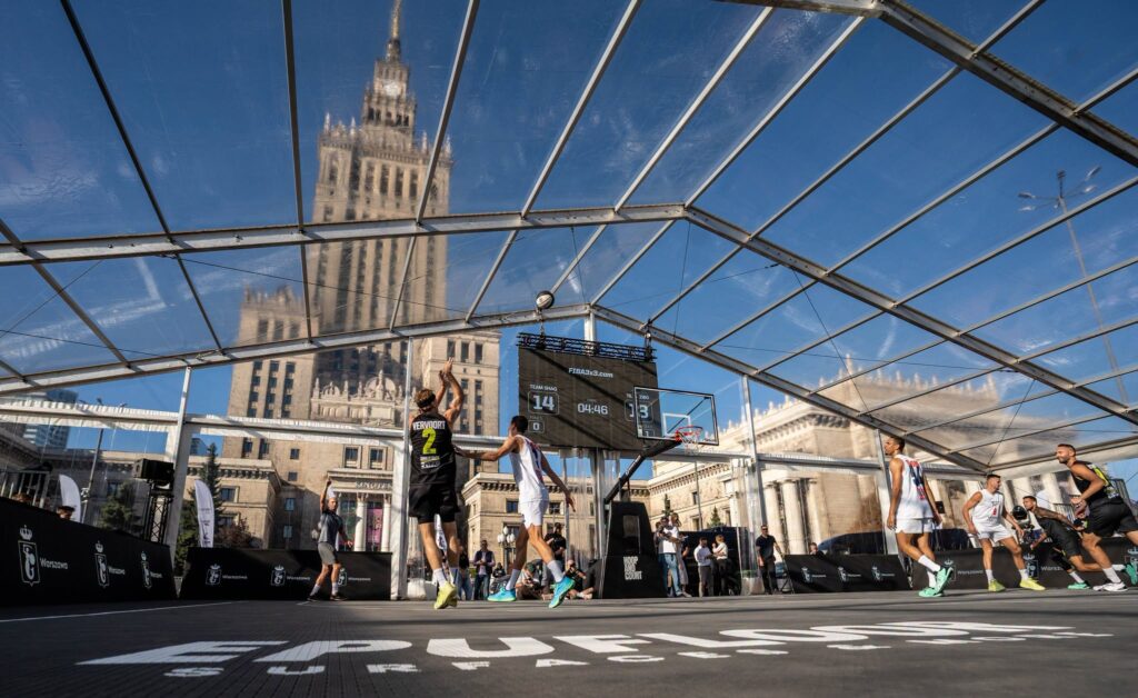 Koszykarze rozgrywają mecz na tymczasowym boisku pod przeszkloną konstrukcją, z Pałacem Kultury i Nauki w tle.