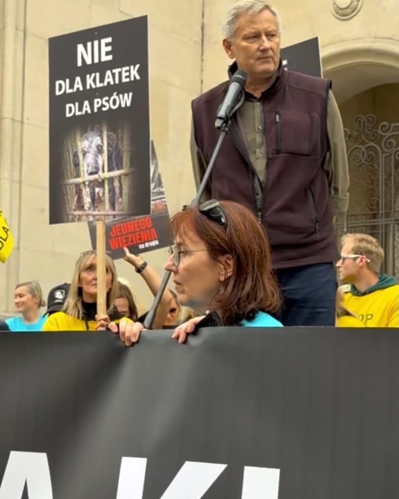 Uczestnicy protestu z transparentem „NIE DLA KLATEK DLA PSÓW”, mężczyzna przemawia przy mikrofonie przed budynkiem.