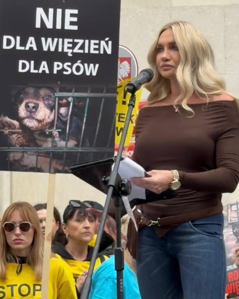Kobieta przemawia przy mikrofonie podczas protestu; obok widoczny plakat z napisem „NIE DLA WIĘZIEŃ DLA PSÓW