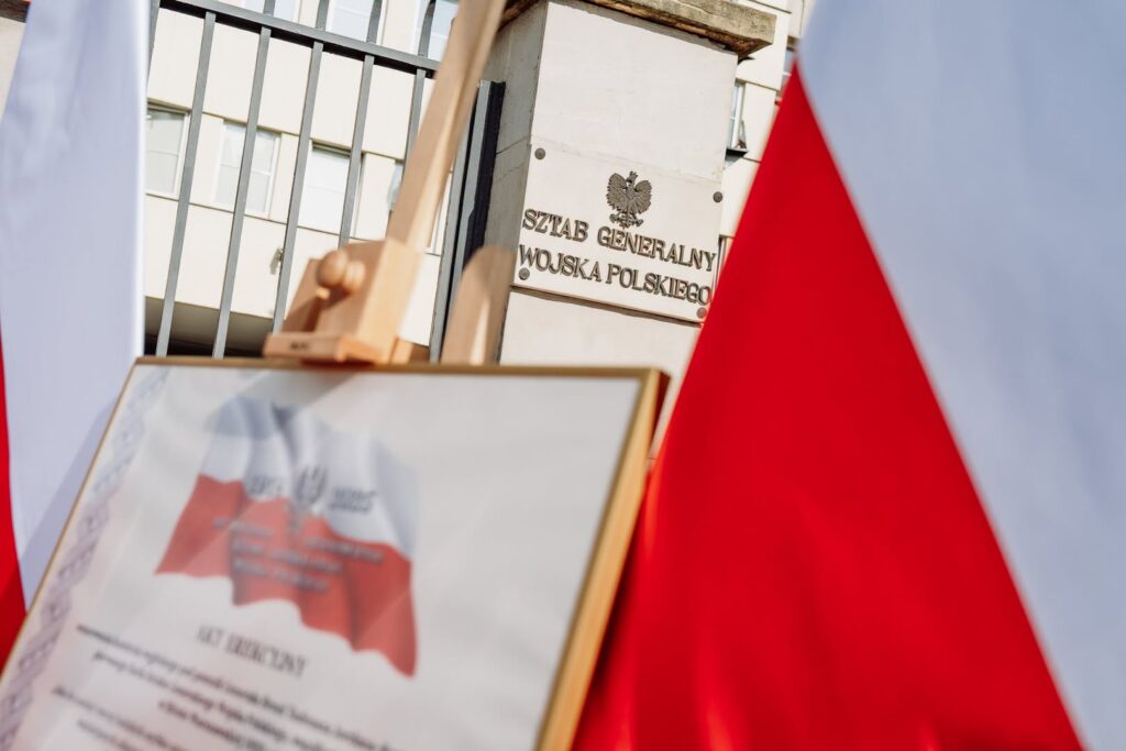 Tablica z napisem „Sztab Generalny Wojska Polskiego” i oprawiony dokument na tle biało-czerwonych flag.
