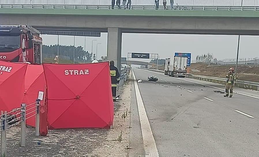 Strażacy zabezpieczają miejsce wypadku na autostradzie pod wiaduktem; na jezdni leży rozbity motocykl.