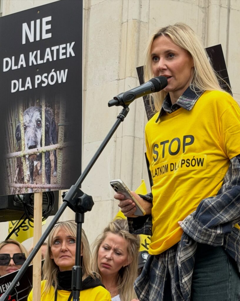 Kobieta w żółtej koszulce przemawia przez mikrofon podczas protestu; obok widoczny transparent „NIE DLA KLATEK DLA PSÓW”.