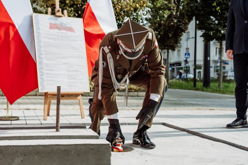 Żołnierz w mundurze kładzie kwiaty przy kamiennej płycie, obok polskich flag i tablicy informacyjnej.