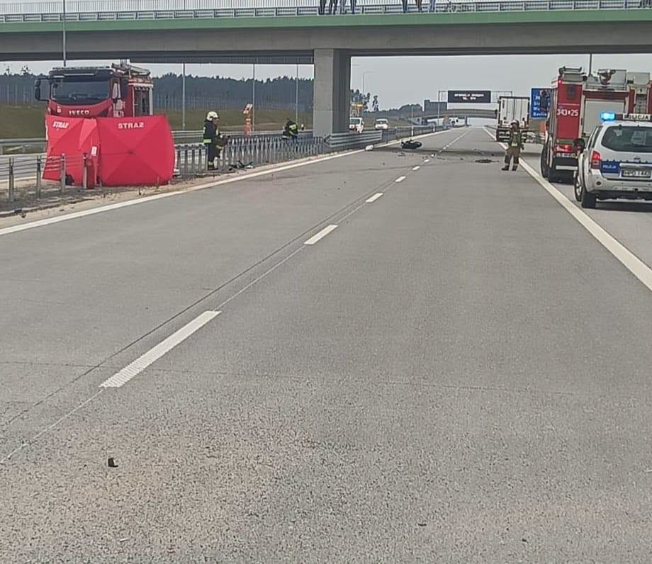 Strażacy i policjanci zabezpieczają miejsce zdarzenia na autostradzie pod wiaduktem, obok stoją wozy służb ratunkowych.