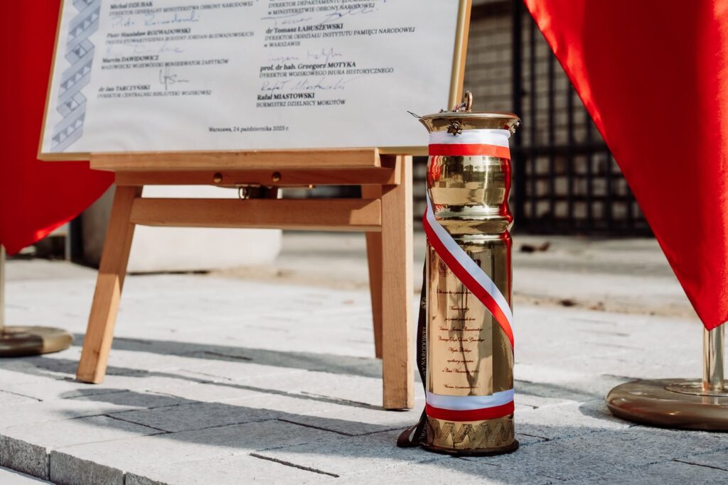 Złota kapsuła czasu przewiązana biało-czerwoną wstęgą stoi obok tablicy na sztaludze, między czerwonymi zasłonami.