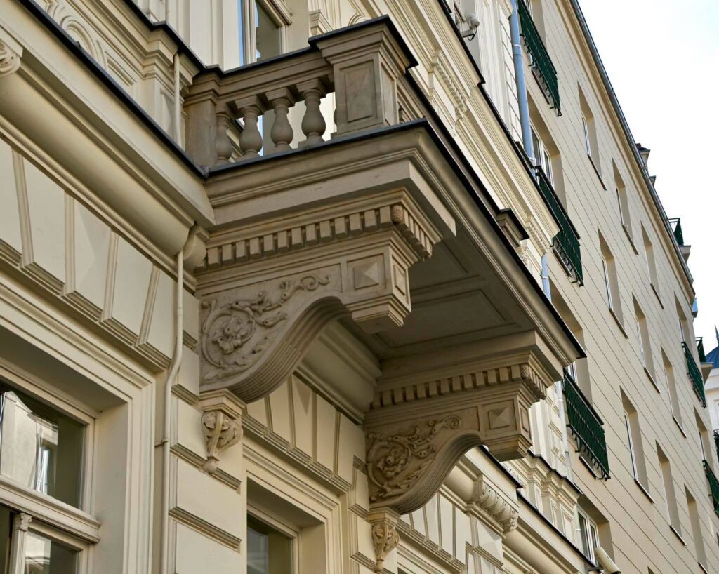 Ozdobny balkon z kamienną balustradą na fasadzie jasnej, historycznej kamienicy z zielonymi okiennicami.