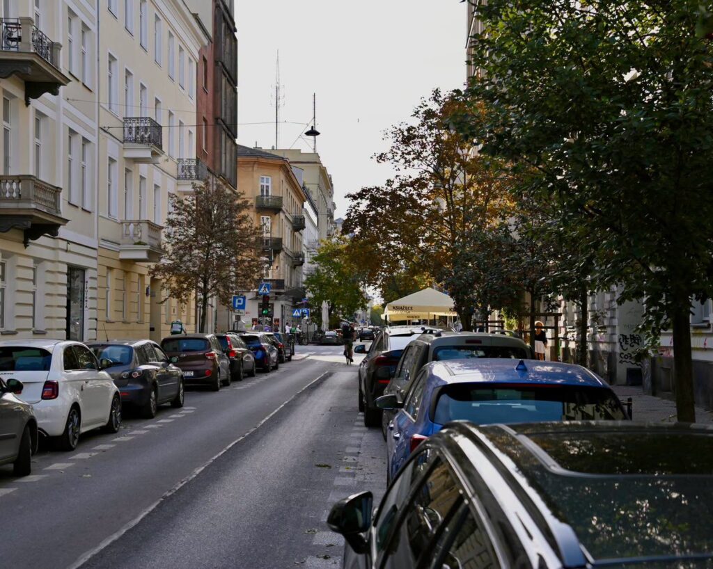 Samochody zaparkowane wzdłuż wąskiej ulicy z kamienicami, drzewami i namiotem przy chodniku.